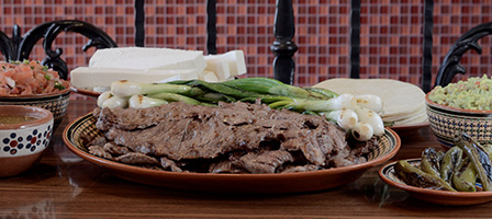 A plate of grilled meat with green onions, surrounded by tortillas, cheese, and salsa
