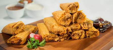 A stack of tamales on a wooden board with a radish, cilantro, and dried chili peppers nearby.