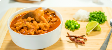 A bowl of stew surrounded by chopped onions, chiles, a lime wedge, and dried red chilies.