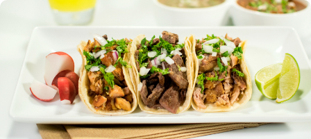 Three tacos with meat, onions, and cilantro on a white plate with radishes and lime wedges.